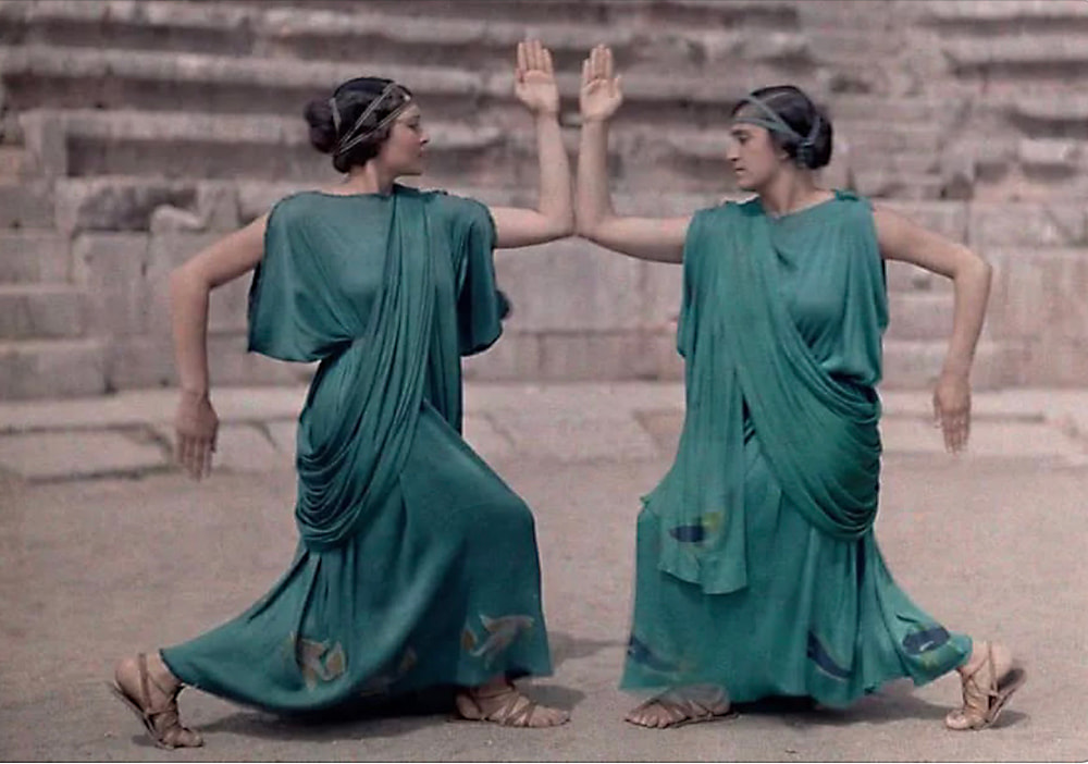 Two women in matching draped garments perform a dance.