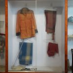 display case with a jacket, pair of pants, and a red garment on hangers, alongside a folded blue-striped blanket