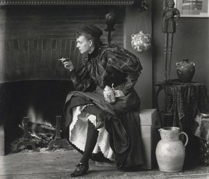 Photo: Frances Benjamin Johnston, full-length portrait, seated in front of fireplace, facing left, holding cigarette in one hand and a beer stein in the other, in her Washington, D.C. studio