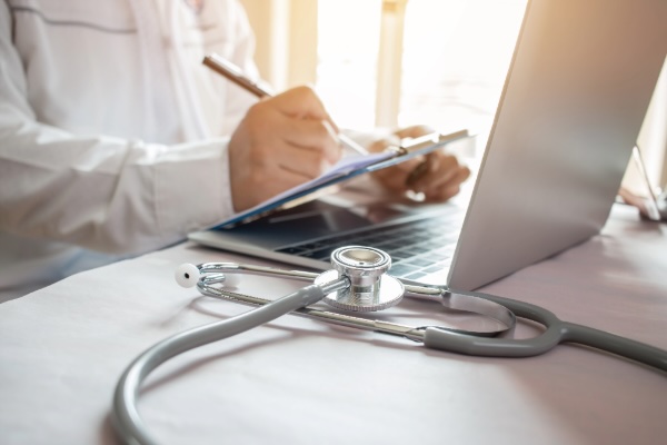 photo of person in a lab coat writing in front of a laptop and stethoscope