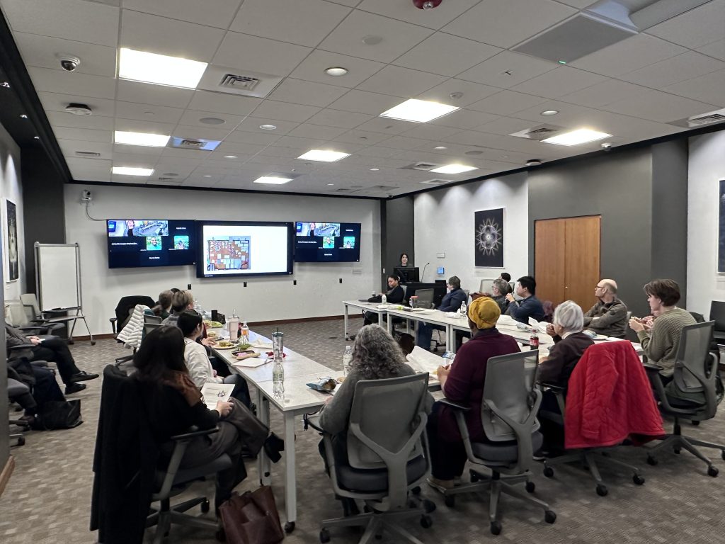 people sit around a large U-shaped table watching a speaker at a podium next to three monitors showing slides and attendees on Zoom