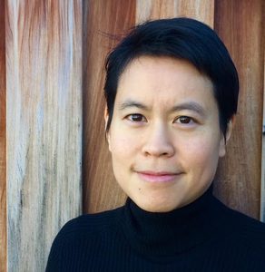 photo of person with short black hair wearing a black turtleneck in front of a a background of vertical wood boards