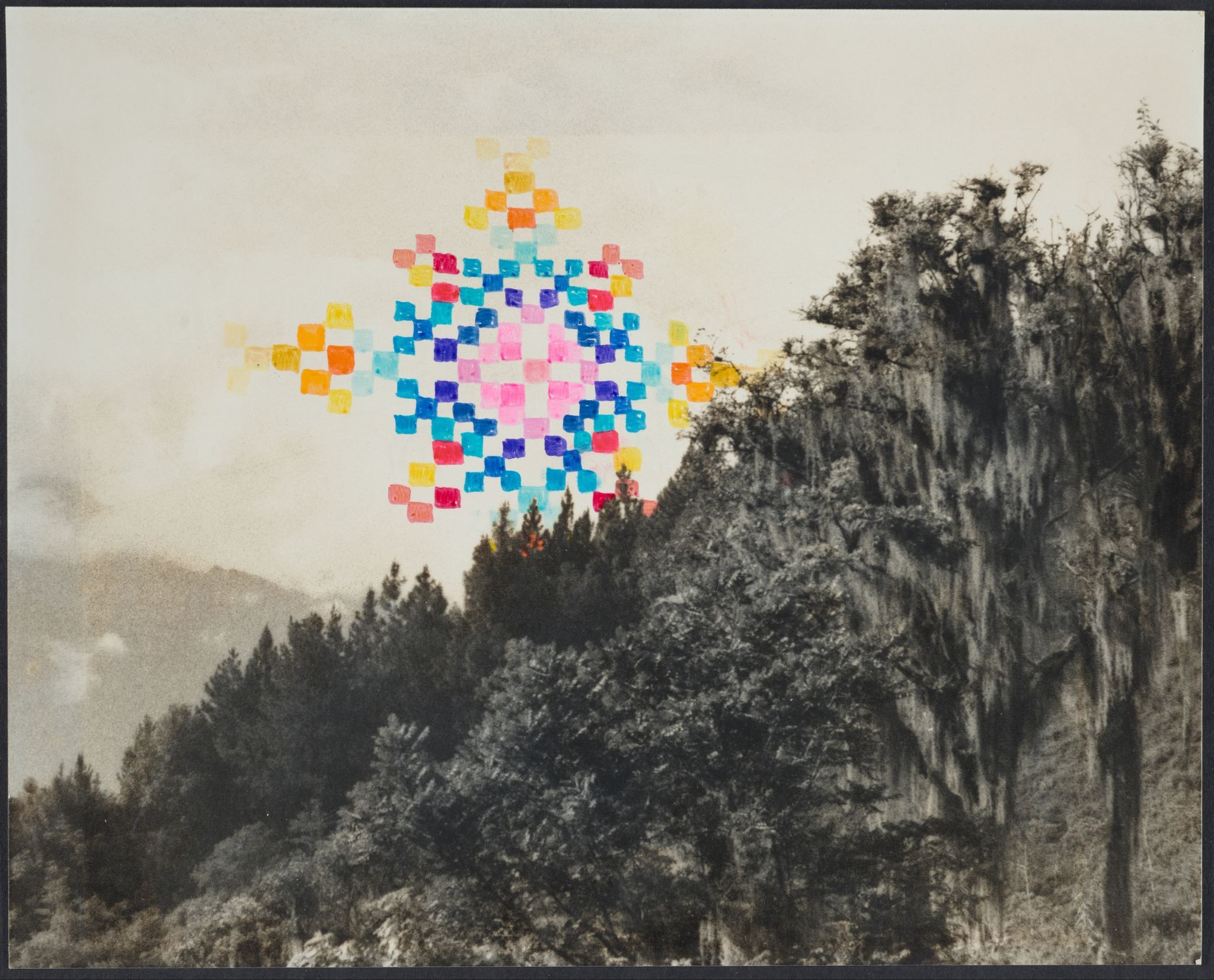 Ya viene Black and white photograph of a dense, forested hillside. A large, brightly colored, symmetrical starburst or mandala design, made of small square pixels, is superimposed over the sky in the upper center.