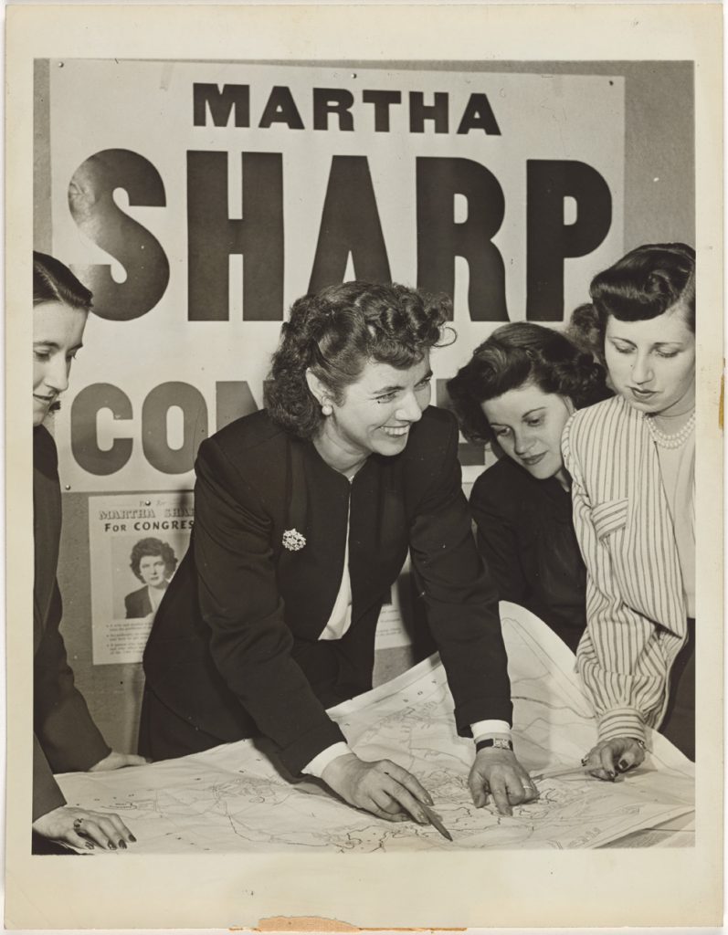 black and white photo of Martha Sharp and women campaign staff in front of campaign poster