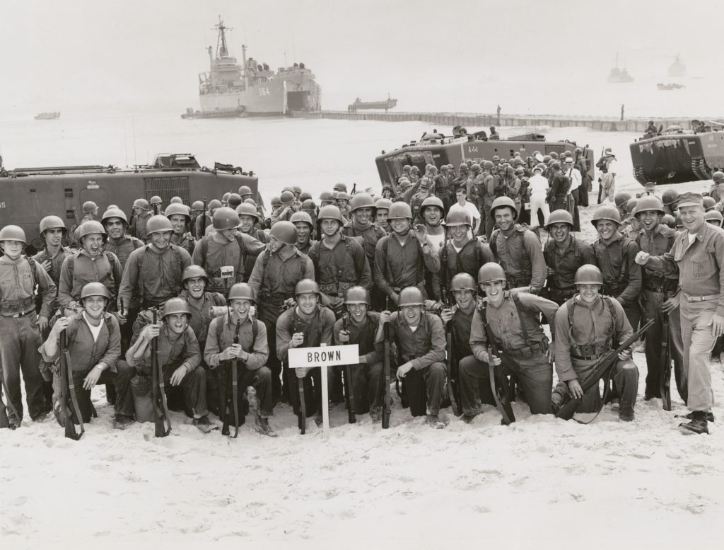 A group of soldiers in front of a transport ship, with a sign saying "Brown" in front of them. Little Creek, VA.